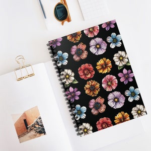 Può includere: Un taccuino a spirale con una copertina nera ornata da un motivo di fiori colorati ad acquerello. I fiori sono nelle tonalità del rosa, viola, blu, arancione e bianco. Il taccuino è aperto su una superficie bianca.