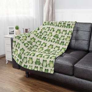 May include: A soft, cream-colored throw blanket with a repeating pattern of green frogs. The blanket is draped over a dark green couch.