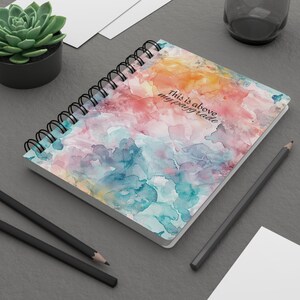 May include: A spiral bound notebook with a watercolor design in shades of blue, pink, and yellow. The cover features the text "This is above my paygrade".