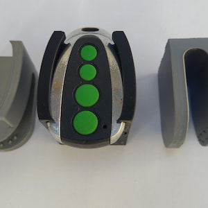 Puede incluir: Un mando a distancia negro y plateado con cinco botones verdes. El mando está sujeto a un soporte de plástico gris.