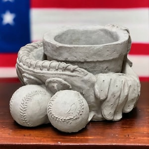 May include: A gray concrete planter shaped like a baseball glove holding two baseballs. The planter has a textured surface and is designed to hold a small plant or flowers.