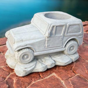 May include: A light gray, concrete planter shaped like a vintage off-road vehicle. The planter has a circular opening on top and sits on a rocky base. The vehicle has detailed tires and windows.