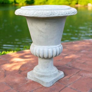 May include: A light gray, ornate garden urn with a wide, decorative rim and a fluted midsection. The urn sits on a square base and is made of a stone-like material. The background features a blurred view of a body of water.