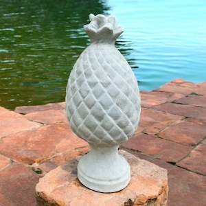 May include: A light gray, textured pineapple statue with a detailed diamond pattern. The pineapple sits on a pedestal base and is displayed outdoors on a stone surface, with a body of water in the background. The statue is a decorative garden accent.