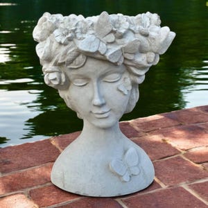 May include: A gray stone planter shaped like a woman's head and shoulders. The woman has closed eyes and a serene expression. The planter is adorned with floral and butterfly details around the head and on the chest.