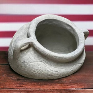 Peut inclure: Un pot en céramique gris texturé avec deux poignées et une large ouverture. Le pot est posé sur une surface en bois avec un drapeau américain en arrière-plan. Le design du pot comprend des détails subtils sculptés.