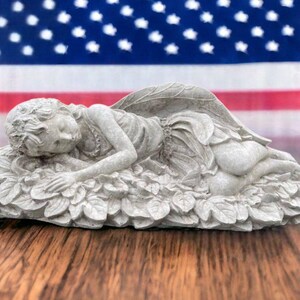 May include: A white concrete garden statue of a sleeping fairy with wings lying on a bed of leaves.