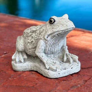 May include: A white ceramic frog statue with black eyes, sitting on a stone base. The frog is positioned on a red brick surface.