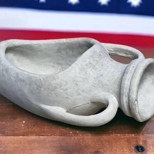May include: A gray ceramic planter shaped like an amphora, a traditional Greek wine jar, with a wide opening at the top and two handles on the sides. The planter is sitting on a wooden surface.
