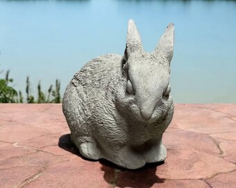 May include: A gray concrete rabbit statue with a realistic design. The rabbit is sitting with its head slightly tilted down and its eyes closed.