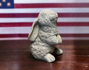 May include: A gray concrete rabbit statue with a textured surface. The rabbit is standing on its hind legs with its front paws clasped together. The rabbit is looking up.