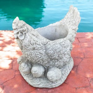 May include: A gray, textured chicken-shaped planter. The planter has a detailed design, including a comb, wattle, and feather details. The base features three rounded feet. The planter is made of a durable material and is suitable for outdoor use.
