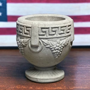 May include: A small, grey, decorative urn with a classic design. The urn features a Greek key pattern around the top and grape clusters on the body. It has a pedestal base and two handles. The background includes an American flag.