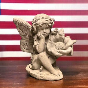 May include: A small, gray, concrete fairy figurine with wings and a flower. The fairy is sitting with her head resting on her hand, looking down at the flower.