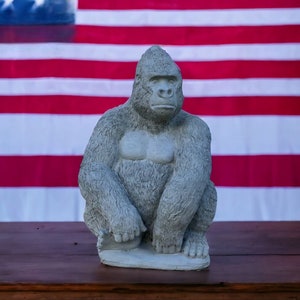 May include: A gray concrete gorilla statue sitting in front of an American flag. The gorilla is sitting with its legs crossed and its arms resting on its knees.