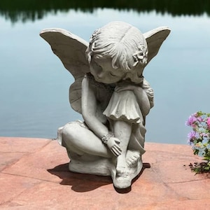 May include: A gray stone sculpture of a fairy with wings sitting with her head resting on her arm. The fairy is wearing a dress and has flowers in her hair.