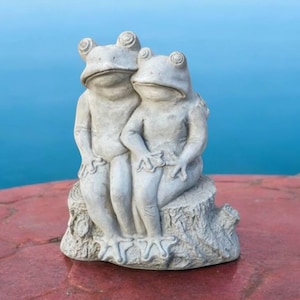 May include: A pair of gray concrete frog figurines sitting on a tree stump. The frogs are holding hands and have a sweet expression on their faces.