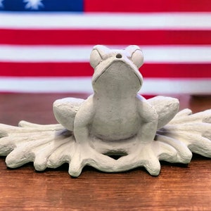 May include: A white ceramic frog figurine sitting on a leaf-shaped base. The frog is looking up with its mouth open. The background is a blurred image of an American flag.