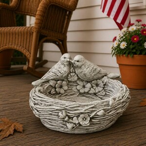 May include: A gray stone bird bath featuring two birds perched on the rim of a nest-shaped basin. The exterior of the basin is textured to resemble woven twigs and is decorated with small sculpted flowers around the circumference of the bowl.