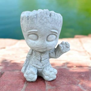May include: A small, gray concrete statue of a character with a tree-like appearance. The statue has a friendly expression and is waving with one hand.