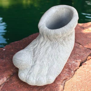 May include: A gray concrete planter shaped like a large foot. The foot has detailed toes, fur-like texture, and an open top for planting. The planter is sitting on a red brick surface with a green water background.