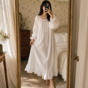 May include: A long-sleeved white nightgown featuring a square neckline with delicate lace trim, a button-down front placket, gathered fabric at the bodice, and a ruffled hemline. The garment is made of a lightweight, opaque fabric with a soft texture.