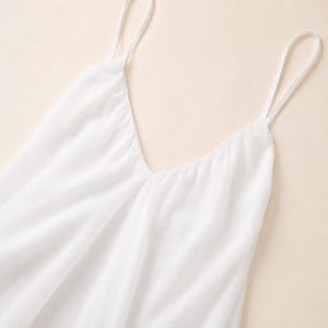 Puede incluir: Una camiseta de tirantes blanca lisa con un escote en V profundo y tirantes finos. La prenda est&aacute; confeccionada con un tejido ligero y semitransparente que cae con suavidad, creando pliegues delicados y una silueta fluida en el torso.