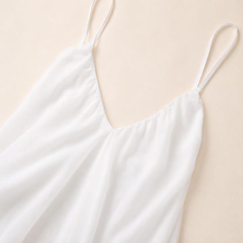 Puede incluir: Una camiseta de tirantes blanca lisa con un escote en V profundo y tirantes finos. La prenda est&aacute; confeccionada con un tejido ligero y semitransparente que cae con suavidad, creando pliegues delicados y una silueta fluida en el torso.