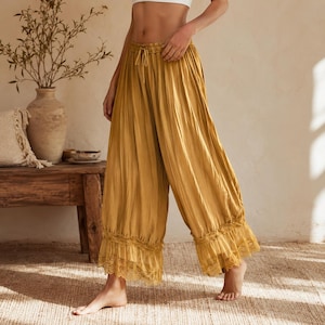 Puede incluir: Pantalones anchos de color amarillo mostaza con textura arrugada, cintura con cord&oacute;n y un dobladillo de doble capa con detalles de encaje intrincado. Los pantalones de corte holgado llegan a una longitud recortada, mostrando una silueta fluida.
