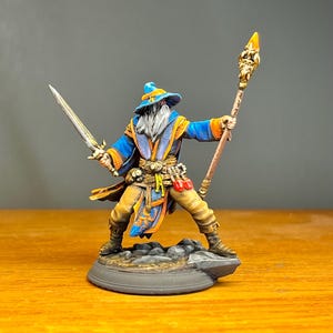 May include: A miniature figurine of a wizard with a long white beard, wearing a blue hat and robe with orange trim. The figure holds a silver sword in the right hand and a tall staff with a glowing yellow tip in the left hand, standing on a base.