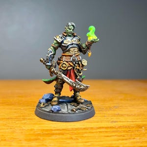 May include: A miniature fantasy figure of a warrior with green skin, holding two swords and a glowing green orb. The figure is clad in detailed armor with gold and silver accents, standing on a stone base. The figure is posed in a dynamic stance.