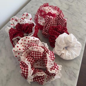 Könnte beinhalten: Ein Set aus fünf roten und weißen Haargummis mit verschiedenen Mustern, darunter Gingham, Punkte und Kirschen. Die Haargummis sind aus Stoff und haben einen Spitzenbesatz.