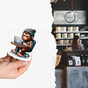 May include: A sticker featuring a cartoon illustration of a brown and orange Bigfoot wearing a brown and white plaid shirt and a blue beanie hat. The Bigfoot is sitting at a desk with a laptop computer and typing on the keyboard.