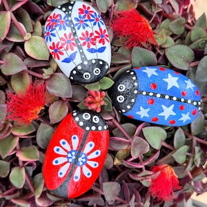 May include: Hand-painted decorative rocks in the shape of ladybugs. The rocks are painted in red, white, and blue with floral and star designs. The rocks are set among green and purple foliage with red flowers.