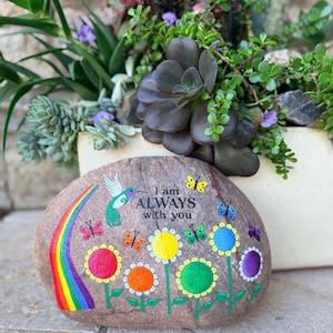 May include: A painted rock with a rainbow, hummingbird, and colorful flowers. The text "I am ALWAYS with you" is displayed. Butterflies are scattered around the design. The rock is placed in front of a planter with succulents and other plants.