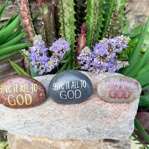 May include: Three smooth, rounded river stones arranged in a row. Each stone features the text "GIVE IT ALL TO GOD" painted in a different color: yellow on a brown stone, white on a dark gray stone, and red on a light gray stone.