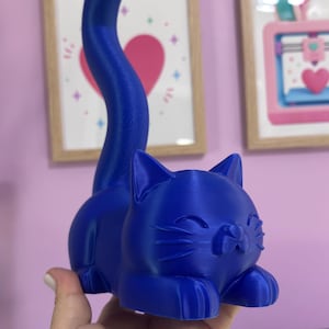 May include: A solid blue cat figurine with a smooth finish, featuring a long, upright tail and a resting posture. The cat has pointed ears, closed eyes, and visible whiskers, with a distinct layered texture characteristic of a 3D printed object.