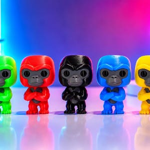 K&ouml;nnte beinhalten: F&uuml;nf kleine Gorillafiguren stehen in einer Reihe, jede in einer anderen Volltonfarbe: Neongr&uuml;n, leuchtendes Rot, Schwarz, K&ouml;nigsblau und leuchtendes Gelb. Jede Figur hat ein dunkelgraues Gesicht, gro&szlig;e runde Augen und verschr&auml;nkte Arme.