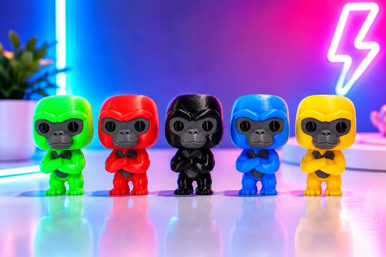 K&ouml;nnte beinhalten: F&uuml;nf kleine Gorillafiguren stehen in einer Reihe, jede in einer anderen Volltonfarbe: Neongr&uuml;n, leuchtendes Rot, Schwarz, K&ouml;nigsblau und leuchtendes Gelb. Jede Figur hat ein dunkelgraues Gesicht, gro&szlig;e runde Augen und verschr&auml;nkte Arme.