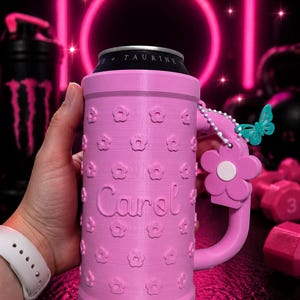 May include: Pink can cooler with a handle and flower details. The name "Carol" is printed on the side. A small butterfly charm hangs from the handle. The cooler is designed to hold a standard beverage can.