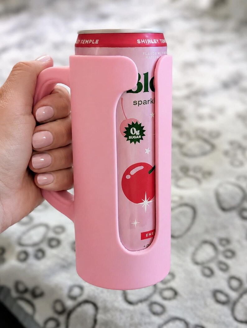 Pu&ograve; includere: Un portabevande rosa con manico contiene una lattina di bevanda frizzante Shirley Temple. La lattina presenta una grafica a ciliegia e le parole "0g Sugar". Il portabevande &egrave; realizzato in materiale morbido.