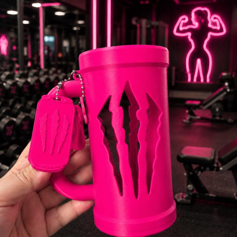 May include: A bright pink cylindrical mug featuring a large cutout of a three-claw scratch logo on the side. A matching pink rectangular tag with a raised three-claw scratch design hangs from the handle by a silver-coloured metal ball chain.