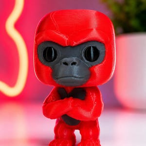 K&ouml;nnte beinhalten: Eine 3D-gedruckte Gorilla-Figur mit leuchtend rotem Kopf, Torso und Gliedma&szlig;en sowie einem dunkelgrauen Gesicht und Brustpanzer. Der Gorilla hat einen gro&szlig;en, abgerundeten Kopf, ausgepr&auml;gte Augenbrauenw&uuml;lste und verschr&auml;nkte Arme vor der Brust.
