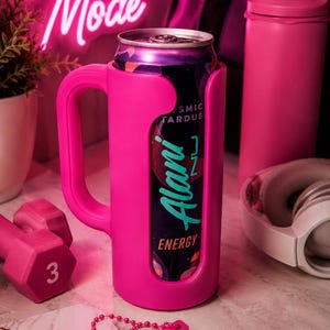 Pu&ograve; includere: Un portabevande rosa acceso con manico, che contiene una lattina di bevanda energetica Alani Nu. L'immagine include un manubrio rosa, una bottiglia d'acqua e delle cuffie, con un'insegna al neon che dice "Beast Mode". &Egrave; visibile anche un taccuino con la parola "FOCUS".