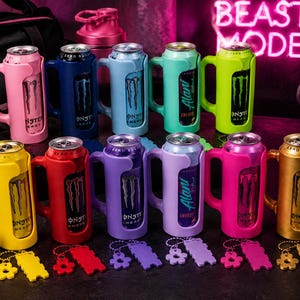 Pu&ograve; includere: Una collezione di colorati porta-lattine Monster Energy con manici. I porta-lattine sono in vari colori, tra cui rosa, blu, verde, giallo, rosso, viola e oro. Ogni porta-lattina ha un portachiavi abbinato. Lo sfondo include attrezzature per l'allenamento e un'insegna al neon che dice "BEAST MODE."