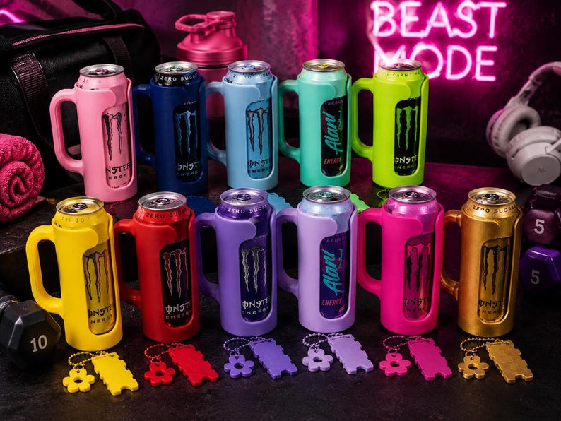 Pu&ograve; includere: Una collezione di colorati porta-lattine Monster Energy con manici. I porta-lattine sono in vari colori, tra cui rosa, blu, verde, giallo, rosso, viola e oro. Ogni porta-lattina ha un portachiavi abbinato. Lo sfondo include attrezzature per l'allenamento e un'insegna al neon che dice "BEAST MODE."