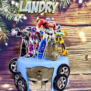 Pode incluir: Um bolo azul decorado com personagens Transformers e um emblema Autobot prateado. O topo do bolo diz "LANDRY" e apresenta o número "4". O bolo é decorado com rodas de carro de brinquedo.