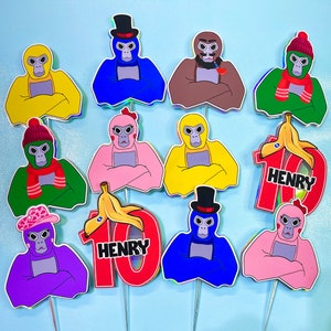May include: Ten colorful cake toppers featuring cartoon monkeys with different expressions and hats. Each topper has a name, "Henry", written on a separate red and white cake topper.