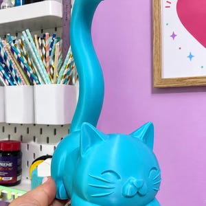 May include: A bright cyan 3D printed cat figurine featuring a long, curved tail extending upward. The cat has a smooth surface, pointed ears, and a simple facial expression with closed eyes and whiskers, designed to hold items on its tail.