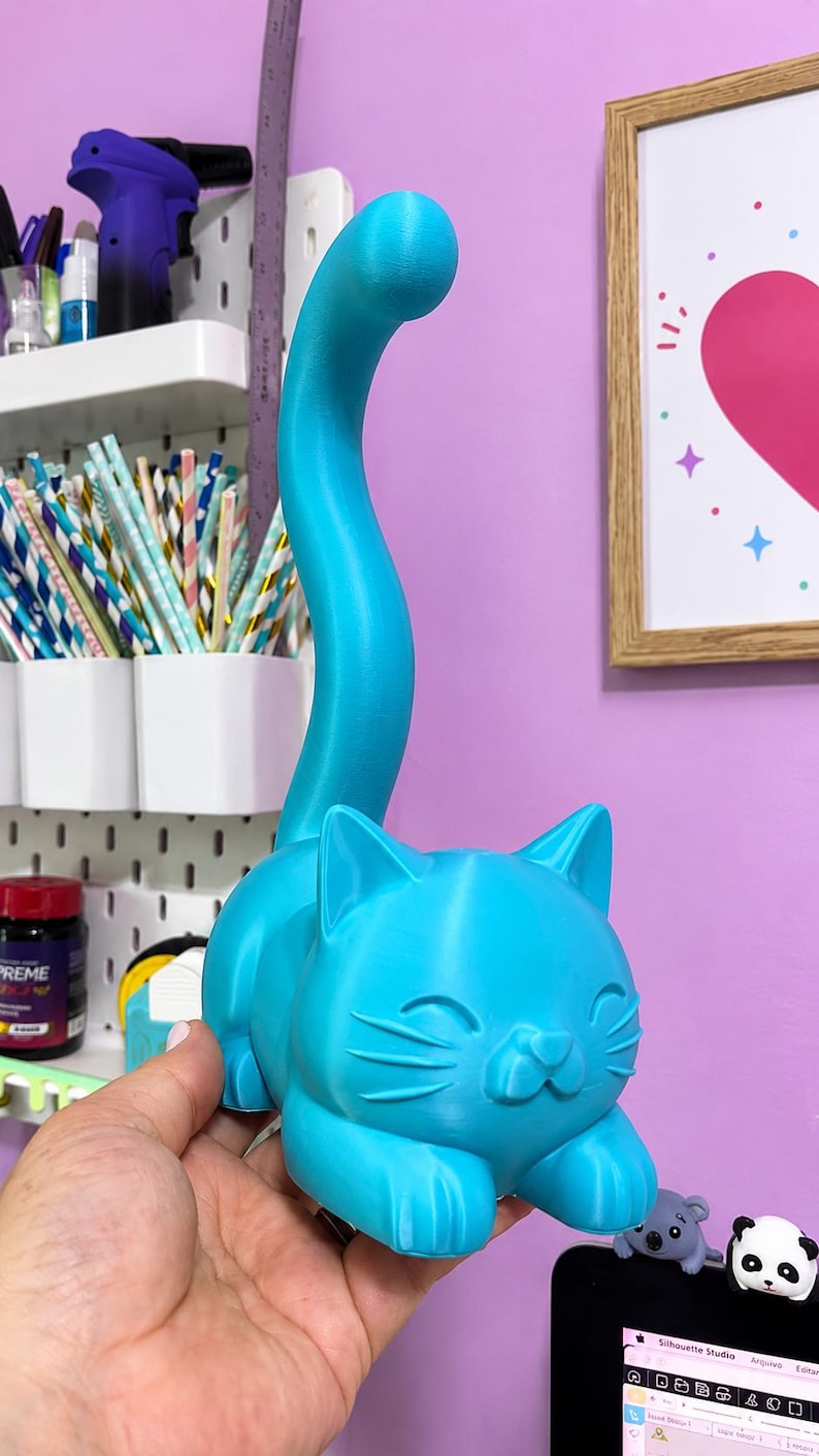 May include: A bright cyan 3D printed cat figurine featuring a long, curved tail extending upward. The cat has a smooth surface, pointed ears, and a simple facial expression with closed eyes and whiskers, designed to hold items on its tail.